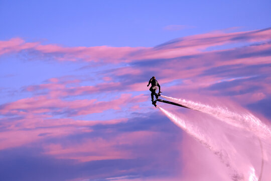 A man in a wetsuit is flying on a flyboard high in the sky. An athlete in water jet boots performs tricks in flight against the background of clouds at sunset. Male doing extreme sports.