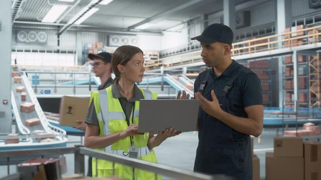Distrubtion Fullfilment Center: Supervisor and Manager Talk, use Laptop Computer. Logistics and Transportation Warehouse Facility with Conveyor Loading Boxes with Products for Delivery to Clients.