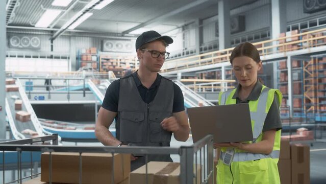 Caucasian Female Logistics Specialist And Male Stocking Associate Talking, Using Laptop In Warehouse Facility With Conveyor Belt. Man Using Barcode Reader To Check Inventory In Distribution Center.
