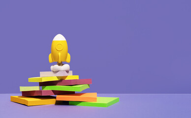 Yellow rocket stands on desk on top of pile of sticky notes summit. purple background. stationery with copy space for back to school or education and creativity concept. Success achievement ideas