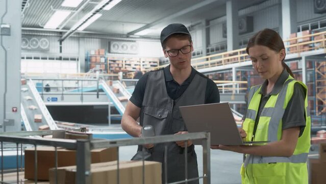 Caucasian Female Logistics Specialist And Male Stocking Associate Talking And Using Laptop In Warehouse Facility With Conveyor Belt. Man Using Barcode Reader To Check Inventory In Sorting Center.