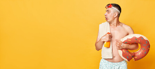 Shocked astonished tourist man at summer resort standing with rubber ring isolated over yellow...