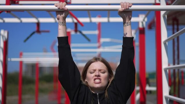 Front view portrait of blond young woman making effort doing pullups in sunbeam outdoors. Motivated Caucasian sportswoman training working out on equipment in slow motion