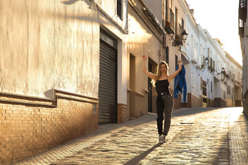 Young woman, beautiful and blonde, wearing a black tank top, blue fur jacket in her hand and jeans, walking down a lonely and lost street during the golden hour. Concept fashion, beauty, sunset.