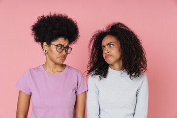 Two upset women looking at each other while standing isolated over pink background