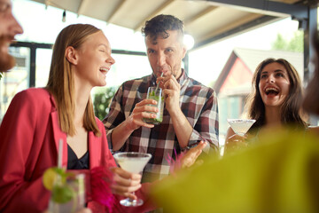 Joyful young people, men and women, friends meeting at cafe, drinking cocktails, talking, having good time together. Concept of friendship, meeting, fun, communication, alcohol drinks