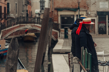 Two gondoliers, dressed in traditional attire, wait by the sidewalk of a Venice canal as they prepare to ferry passengers, with a bridge in the background.