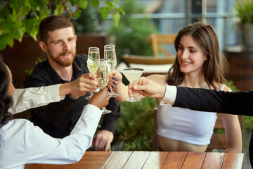 Cheerful young people, employees clinking glasses with cocktails and champagne, celebrating successful project after work in restaurant. Concept of leisure time, business, communication, alcohol drink