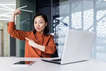 Portrait of a young successful Asian female programmer, developer and IT expert. Sitting at a table and working on a laptop, pointing at the camera with a finger on his hand, a gesture of strength.