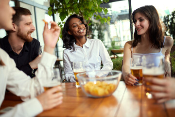 Happy, positive, smiling young people, office workers, colleagues meeting in cafe, pub after work to drink beer, talk and relax. Concept of leisure time, business, communication, fun, alcohol drink