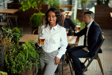 Focus on african young woman, office worker standing with beer. Blurred people on background. Cheerful meeting. Concept of leisure time, business, communication, fun, alcohol drink