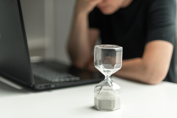 Hourglass in the foreground. A woman is sitting in front of a laptop and is stressed, covering her face with her hands. The concept of lack of time.