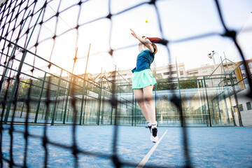 Photo through the net of a paddle tennis court of a female paddle tennis player jumping to hit a...