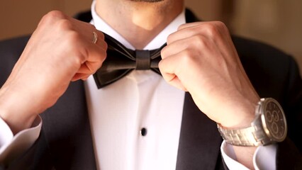 The groom in the black suit adjusts his bow tie of the white shirt - close up