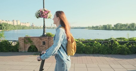Red-haired happy smiling young woman rides an electric scooter in park near river, green trees, following camera, low angle, front side view.