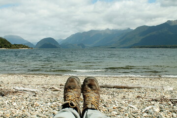 Woman's hiking boots rest on the verge of mountains surrounded lake in midday at Fiordland New...