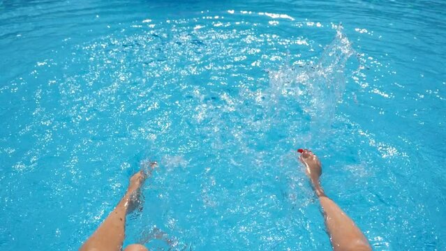 Female legs splashing crystal clear water in pool of hotel. Two friends sitting on edge of basin and enjoying their vacation at sunny day. Concept of summer vacation or holiday. Slow mo Point of view