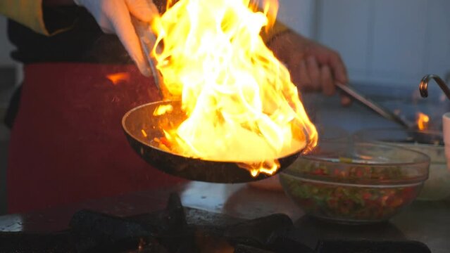 Detail view on flaming skillets in hand of professional chef. Male cook tossing and frying vegetables cooking tasty flambe style dish for visitors of restaurant. Concept of cooking. Slow mo Side view