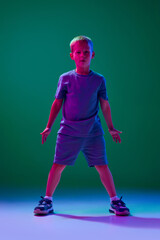 Full-length portrait of little boy, child in casual clothes standing, playing against cyan studio background in neon light. Concept of childhood, lifestyle, emotions, education, fashion, care, ad