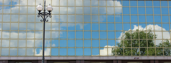 Reflection Of Trees, Sky And Clouds In Glass Facade Of Modern Building With Street Lamp In Front 
