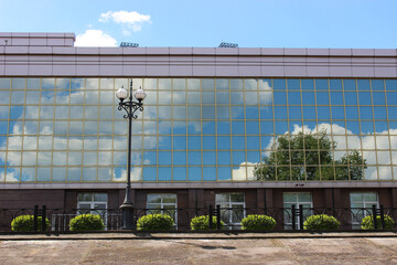 Fototapeta na wymiar Green Tree And Clouds On A Blue Sky Reflects In Mirror Glass Wall Of The Office Building With Green Bushes In Front Of It 