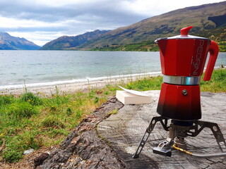 A red coffee maker and a book to start the day in front of the blue lake and green hills during camping