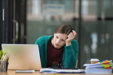 A tired businesswoman under stress works at a laptop while sitting at a table at home and holds her...