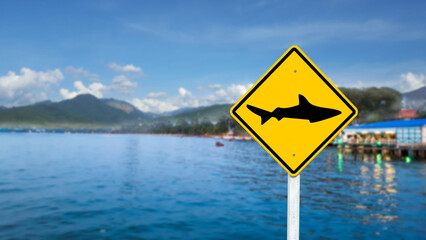 Graphic of shark on yellow sign on blurred sea and restaurant in background to warn people not to...