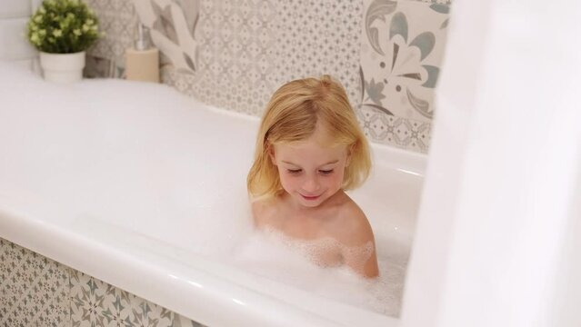 Adorable caucasian three years old gir with curly hair taking bath with white foam bubbles, playing,laughing,having fun.Carefree childhood, kid body care and hygiene concept.