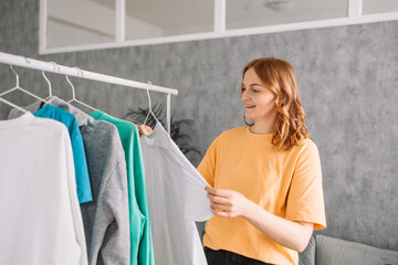 Clothing, purchasing, style concept. Happy blonde haired woman chooses clothes at home wardrobe, holds comfortable jumper on hangers, outfits on racks in background, messages via mobile phone. High