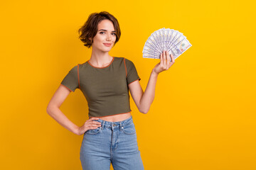 Portrait of positive adorable girl with short hairdo wear khaki t-shirt hold dollars hand on waist isolated on yellow color background