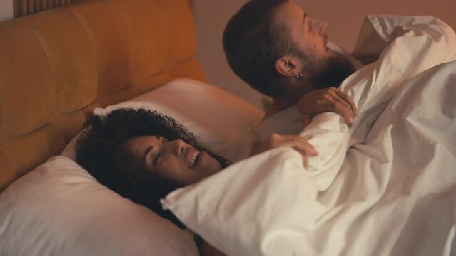 Laughing interracial couple having fun in bed, pulling away blanket, romance