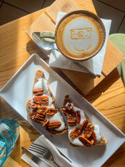 Colazione di un bistrot fotografata dall'alto con cappuccino con una macchina fotografica disegnata sul latte e due toast con noci pecan, yogurt greco e miele - obrazy, fototapety, plakaty