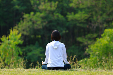 Rear view of woman in hoodie is relaxingly practicing meditation yoga in forest full of grass...