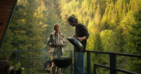 A young couple, a guy and a girl, are relaxing in a country house and frying meat on a special grill against the backdrop of a green coniferous forest