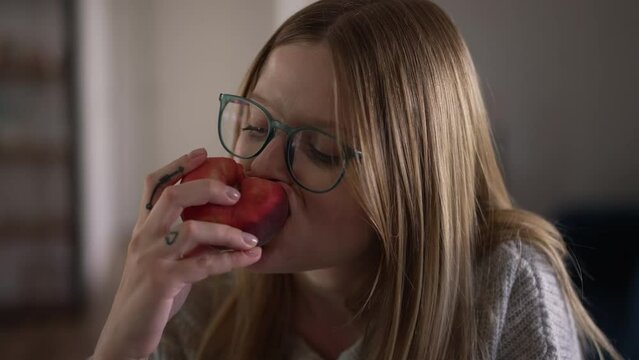 Headshot young woman eating tasty peach in slow motion indoors. Close-up portrait of beautiful Caucasian lady in eyeglasses enjoying delicious vitamin fruit at home