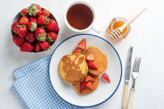 Top view pancakes with strawberries and honey served with cup of black tea. Morning image, breakfast meal on light grey concrete background