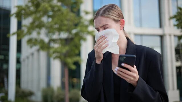 Sick businesswoman holds a tissue and a mobile phone, coughing due to a cold. The young successful woman manager is experiencing allergy symptoms, feeling unwell after catching a cold at work.