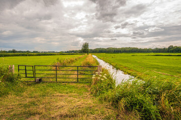 Gate in the foreground of a meadow in a Dutch polder landscape. Along the meadow is a ditch for water drainage. It is a heavily cloudy day in the summer season.
