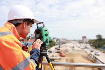 Site young man engineer operating his instrument during roadworks looking at camera. Builder using...
