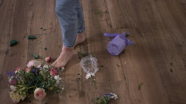 Barefoot legs of young depressed woman walking on wooden floor with broken bouquet kicking flowers leaving in slow motion. High angle view unrecognizable Caucasian sad desperate lady at home indoors