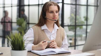 Fototapeta premium Attractive confident business woman, office manager, wearing headset using laptop while making, writing financial repor