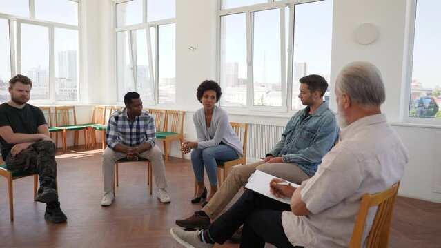 African alcohol addict female sharing anger management issues with people at aa rehab meeting. Stressed young woman with mental health problems talking to patients and psychologist in circle.