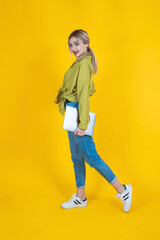 Holding closed laptop, full body side view young blonde woman holding closed laptop. Wearing jeans and green shirt, isolated yellow studio background. Walking, going, IT professional lifestyle concept