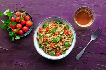 Quinoa tabbouleh salad in a bowl, a healthy dinner with tomatoes and mint, overhead flat lay shot with a drink on a purple background
