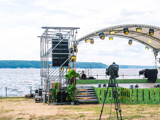 A stage for artists' performances on the riverbank in summer. Concert venue with lighting equipment. Concert on the river bank. A video camera on a tripod in front of the stage.
