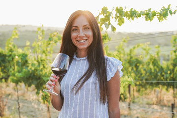 Young woman inside wineyard holding glass of red vine during sunset time and smiling on camera - obrazy, fototapety, plakaty