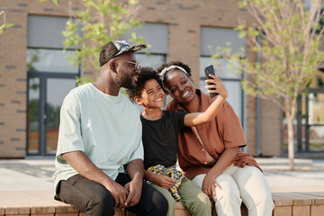 African American family of three making selfie portrait on smartphone while sitting on bench outdoors