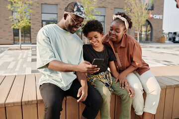 Fototapeta premium African American family sitting on bench and watching photo on smartphone after walk