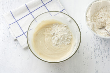 Glass bowl with dough, adding flour on a light blue background, top view. Cooking homemade milk cakes, do it yourself, step by step, step 6